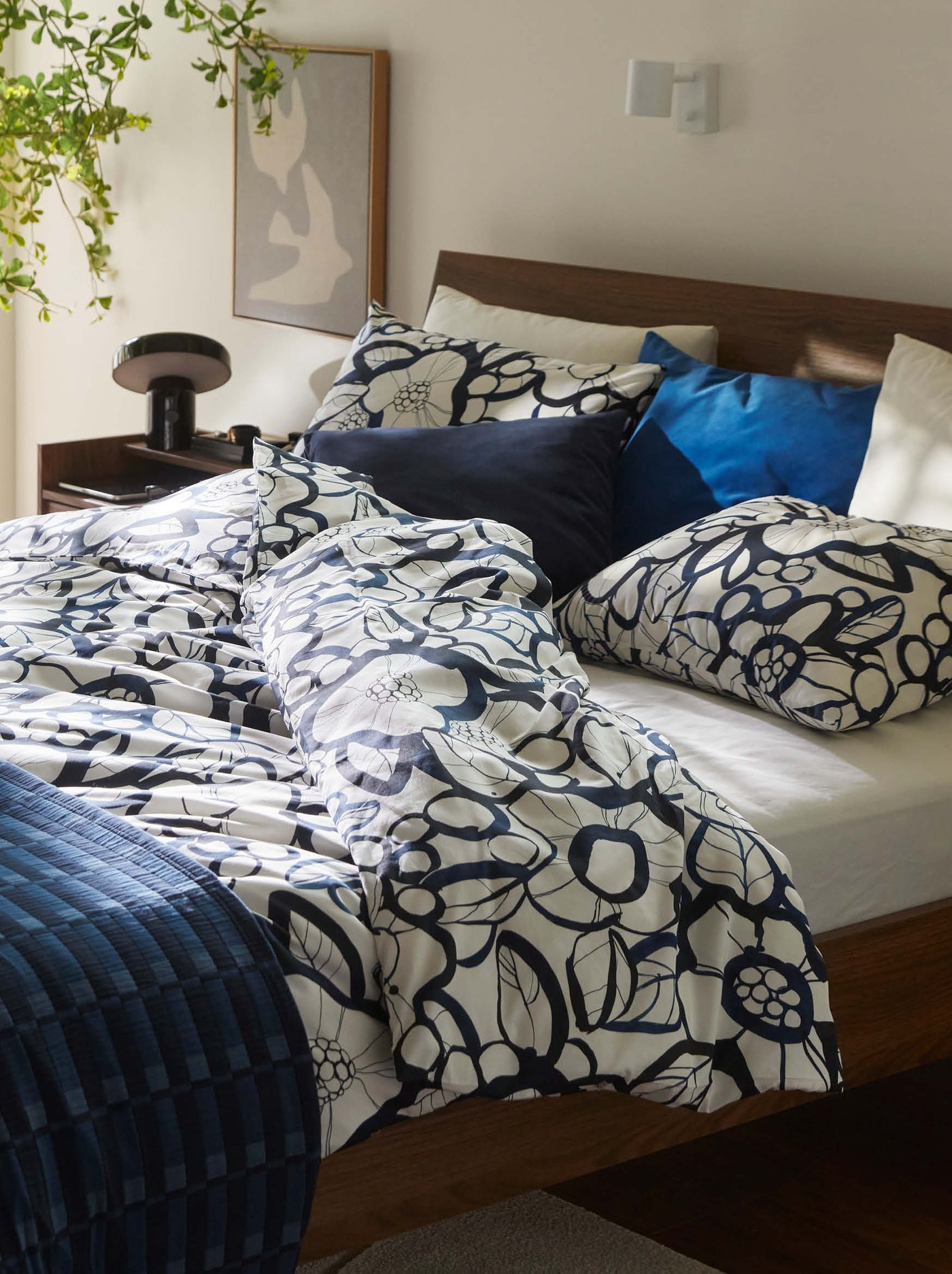 Ein Holzbett mit weiß-marineblauer Bettwäsche mit Blumenmuster, mehreren Kissen, einer Nachttischlampe, Wandbildern und einer hängenden Grünpflanze.