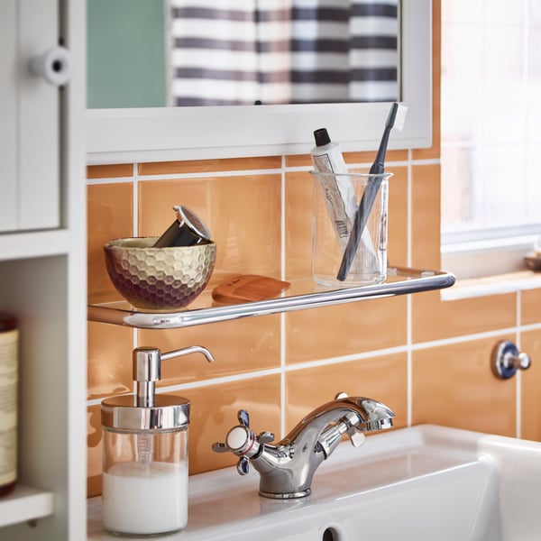 A bathroom sink with RUNSKÄR tap, VOXNAN soap dispenser and VOXNAN glass shelf in an orange tiled bathroom.