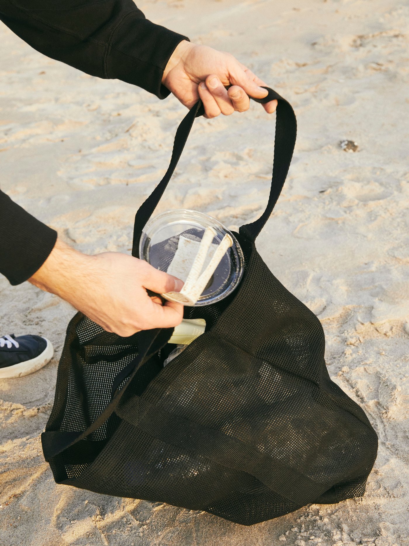 KÅSEBERGA bag in black mesh being used by a person for picking up litter, like plastic waste, on a sandy beach.