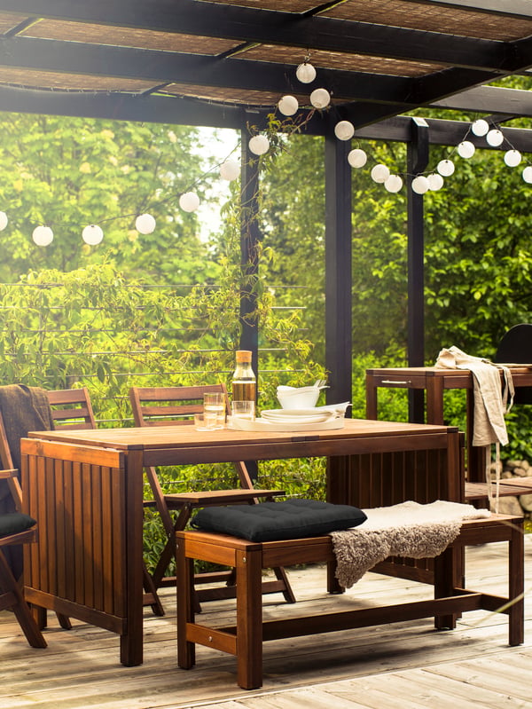 An outdoor table with chairs and a bench with cushions, hanging lighting and green trees in the background.