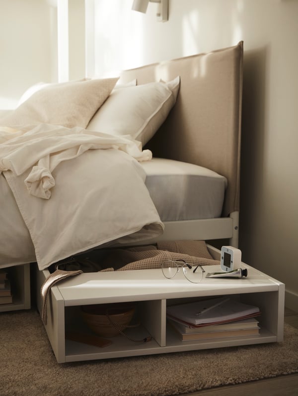 Una mesita de noche/contenedor debajo de la cama FREDVANG blanca junto a una cama KLEPPSTAD blanca/vissle beige vestida con ropa de cama ÄNGSLILJA.