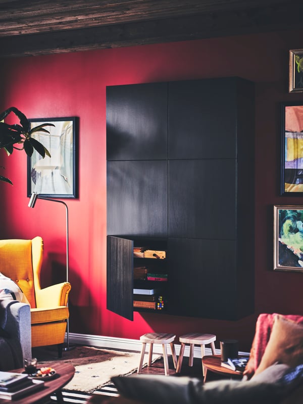 A living room with a storage unit with black-brown LAPPVIKEN doors, with two stools below and a yellow STRANDMON wing chair.