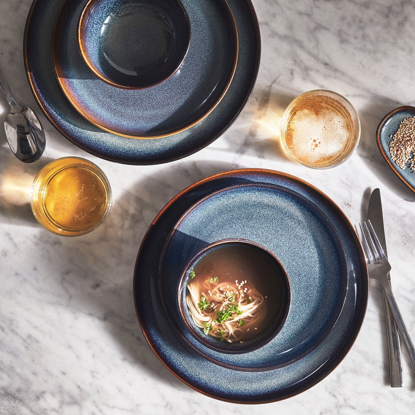 Deux bols GLADELIG bleus dans lesquels se trouvent des aliments variés, déposés sur des assiettes GLADELIG sur une table avec un couteau, une fourchette et une cuillère.