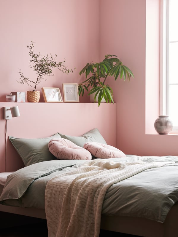Un lit est installé dans le coin d’une chambre rose. Plusieurs plantes d’intérieur et décorations sont posées sur un rebord au-dessus.