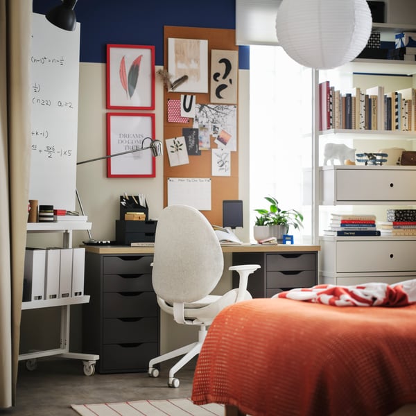 In einem Schlafzimmer steht vor einem ALEX/LAGKAPTEN Schreibtisch ein HATTEFJÄLL Drehstuhl mit Armlehnen in Beige/Weiß. Daneben ist eine Schreib-/Notiztafel zu sehen.