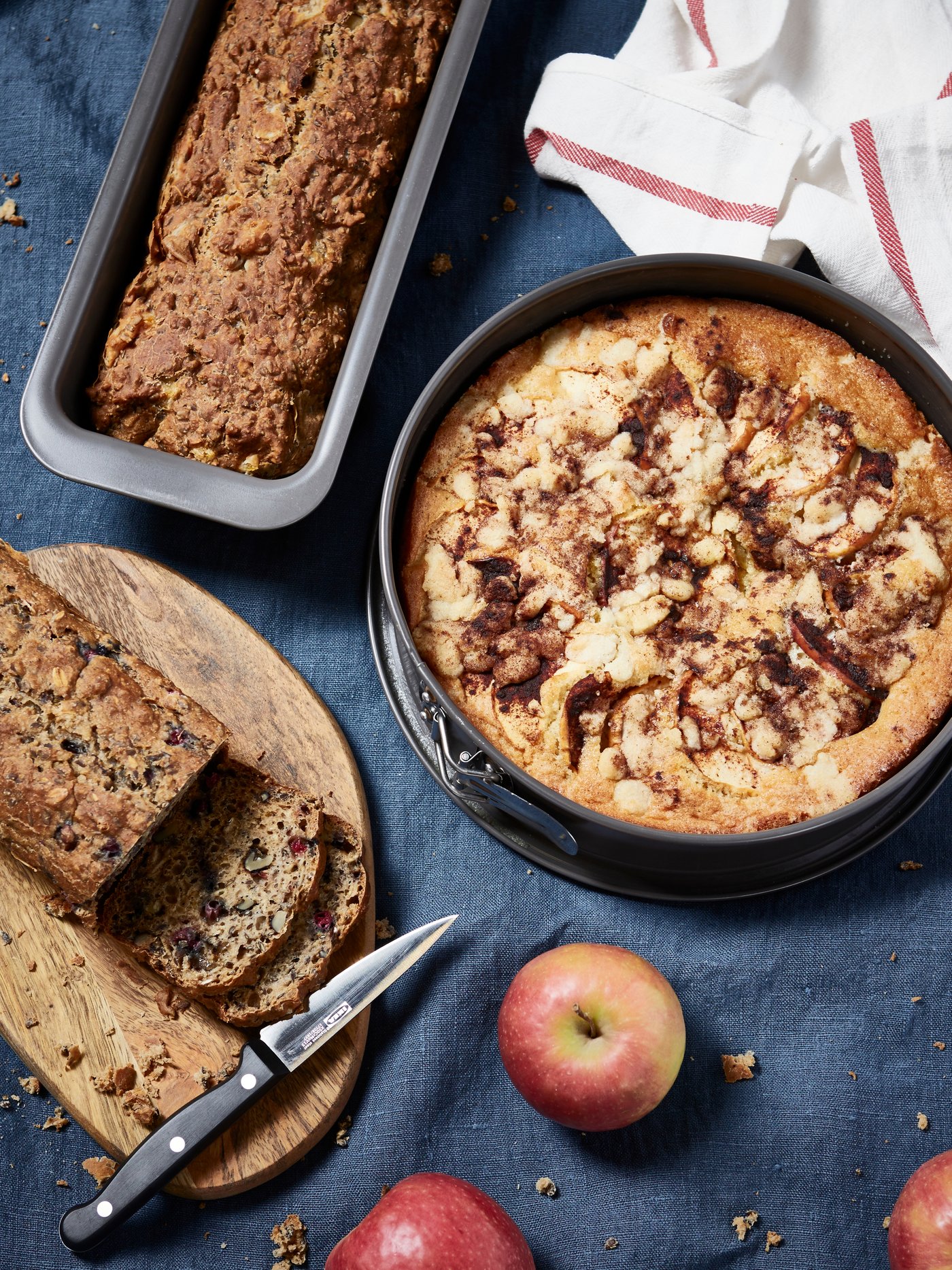 A top view of a HEMMABAK baking form with a cake, next to it there is another HEMMABAK baking form with a bread. There is another bread that is being cut on a cutting board on the table There are also two apples.