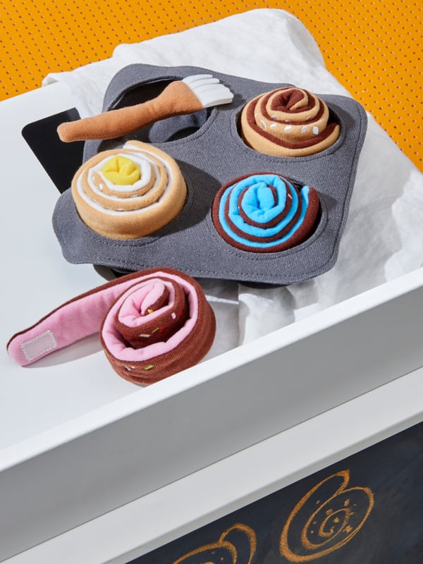 Close-up of DUKTIG toy fabric pastry rolls, pastry brush and grey fabric pan on yellow perforated counter.