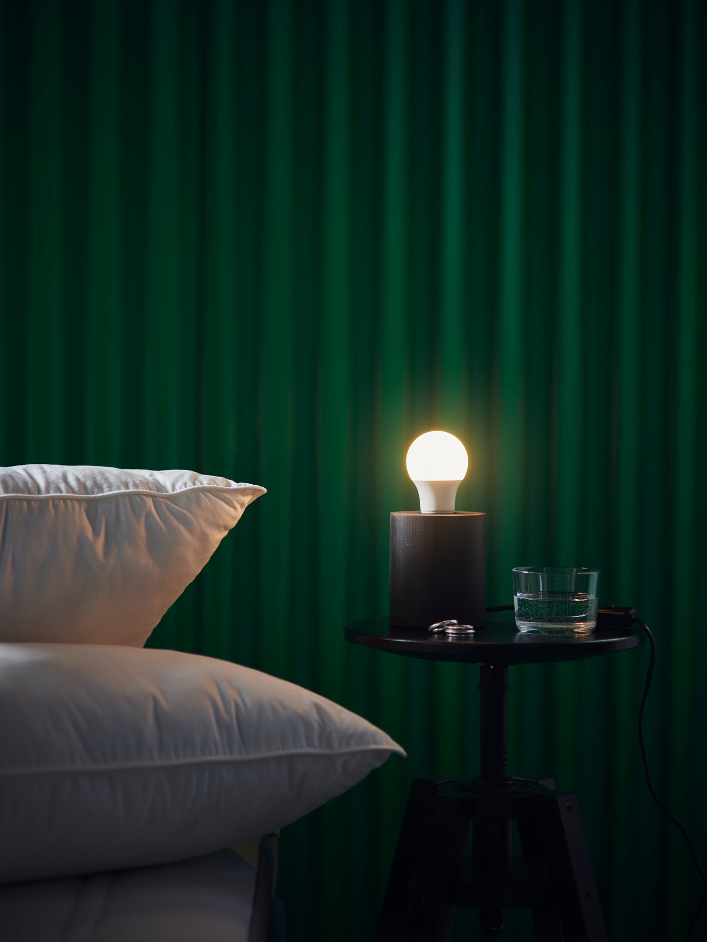 Deux coussins blancs sont empilés à côté d'une petite table noire sur laquelle se trouvent une ampoule SOLHETTA allumée et un verre d'eau. Le mur derrière est vert foncé. 