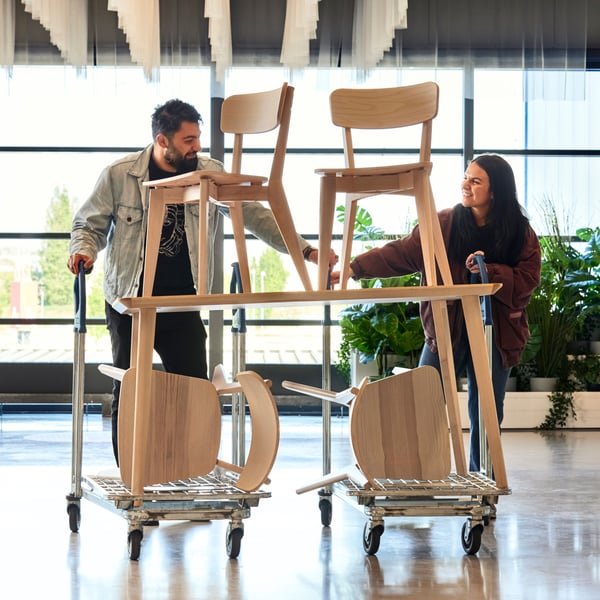 Two people push carts with wooden chairs in a bright space with large windows and indoor plants.