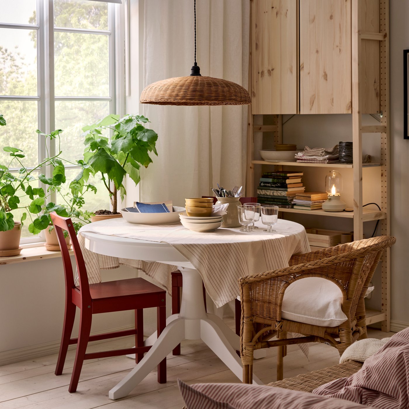A white ROSENTORP table by a window, IVAR shelving unit behind it and different kinds of chairs. Plants on the windowsill.