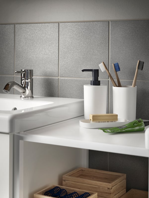 A toothbrush holder with three toothbrushes, a soap dispenser and a tray in black/white stand on a shelf beside a wash-basin.