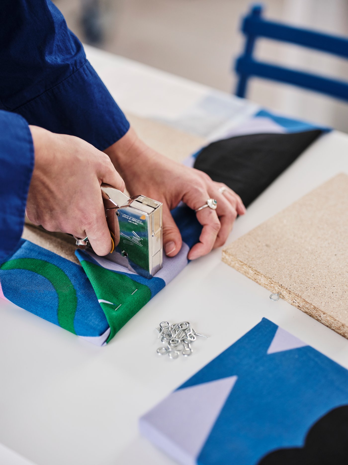 Hands are attaching the FJÄLLDRABA pre-cut fabric in blue, green and lilac onto a wooden board with a staple gun.