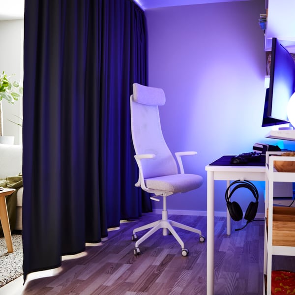 Una silla gamer blanca frente a un escritorio en un espacio con luces encendidas. Una cortina separa el espacio de un living.