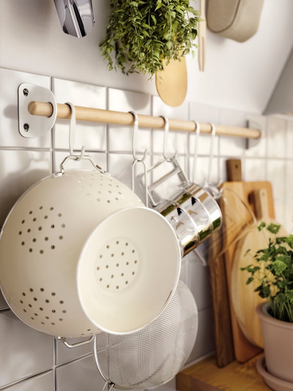Küchenwand mit hängender NEREBY Holzstange zur Aufbewahrung von Küchenutensilien und einem GEMAK Sieb. Kräuterpflanze, Schneidebretter und eine gemütliche, aufgeräumte Atmosphäre.