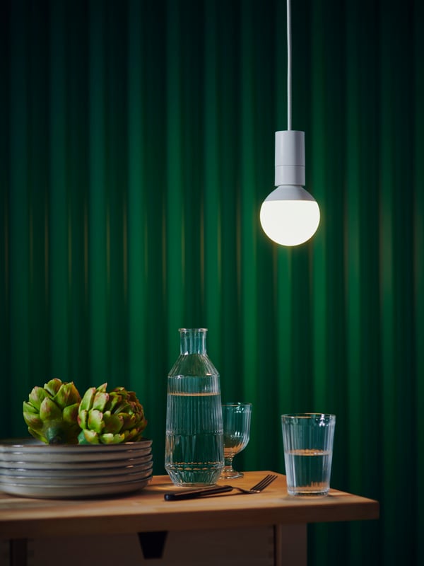 Une table à manger éclairée avec des assiettes, deux artichauts, une carafe d'eau, des verres et des couverts. Le mur derrière est vert foncé. 
