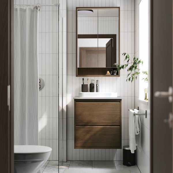 Un meuble lavabo ÄNGSJÖN effet chêne brun avec une armoire à pharmacie au-dessus dans une salle de bains blanche avec fenêtre.
