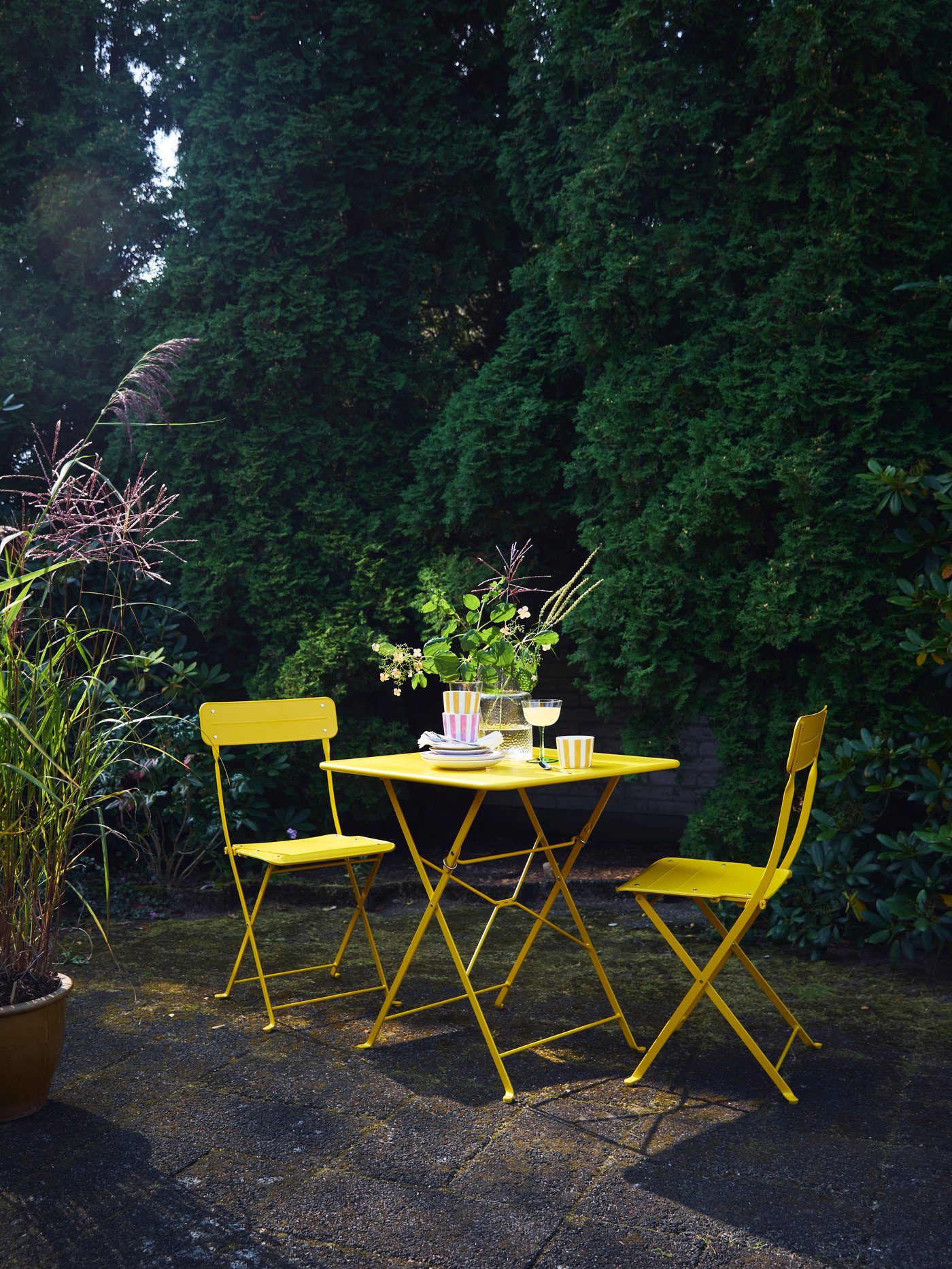Una mesa y dos sillas de exterior de la serie SUNDSÖ en color amarillo claro que ilumina un patio trasero en un ambiente nocturno.