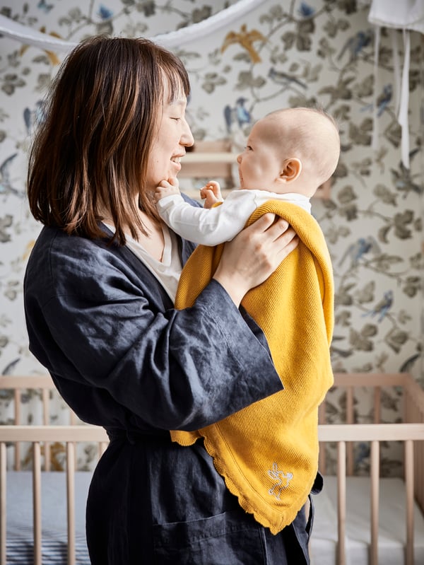 A mom standing in a robe holding her baby wrapped in a yellow blanket.