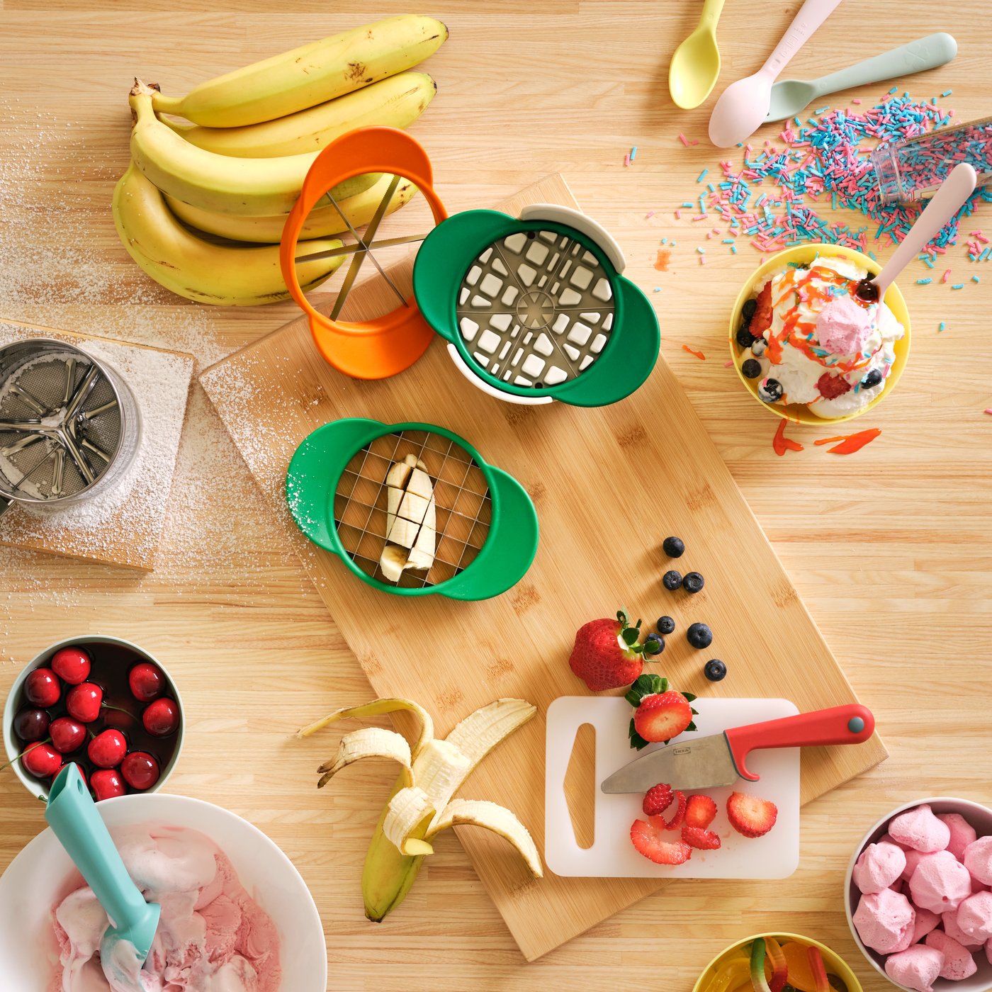 A tabletop with a chopping board holding green and orange UPPFYLLD fruit cutters, surrounded by fruits and bowls of berries.