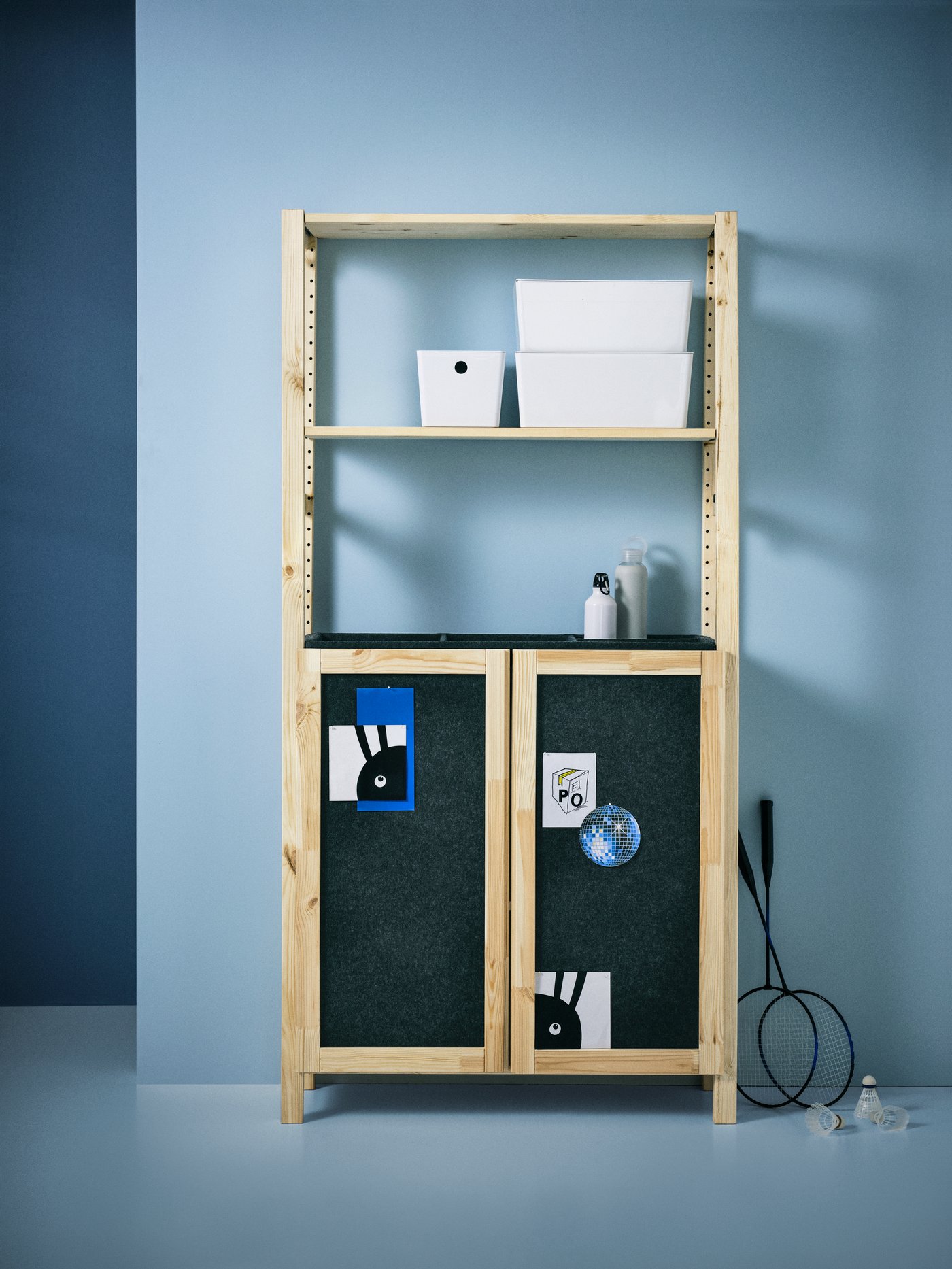 An IVAR shelving unit with two doors, with three white KUGGIS boxes with lids on a shelf, stands against a blue wall.