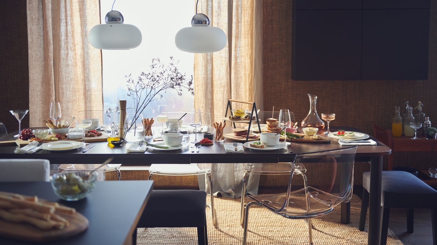 A STRANDTORP extendable dining table in front of a window, laid for a big meal with plates, glasses, flowers and food.