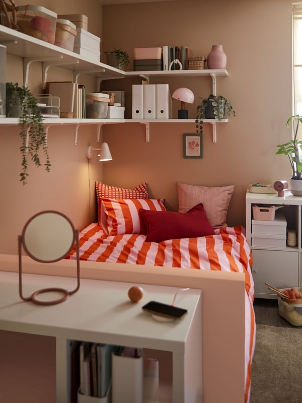 Ein kleines Zimmer mit einem weißen KALLAX-Schreibtisch am Fußende eines Bettes mit Bettwäsche in hellen Farben und Wandregalen darüber.