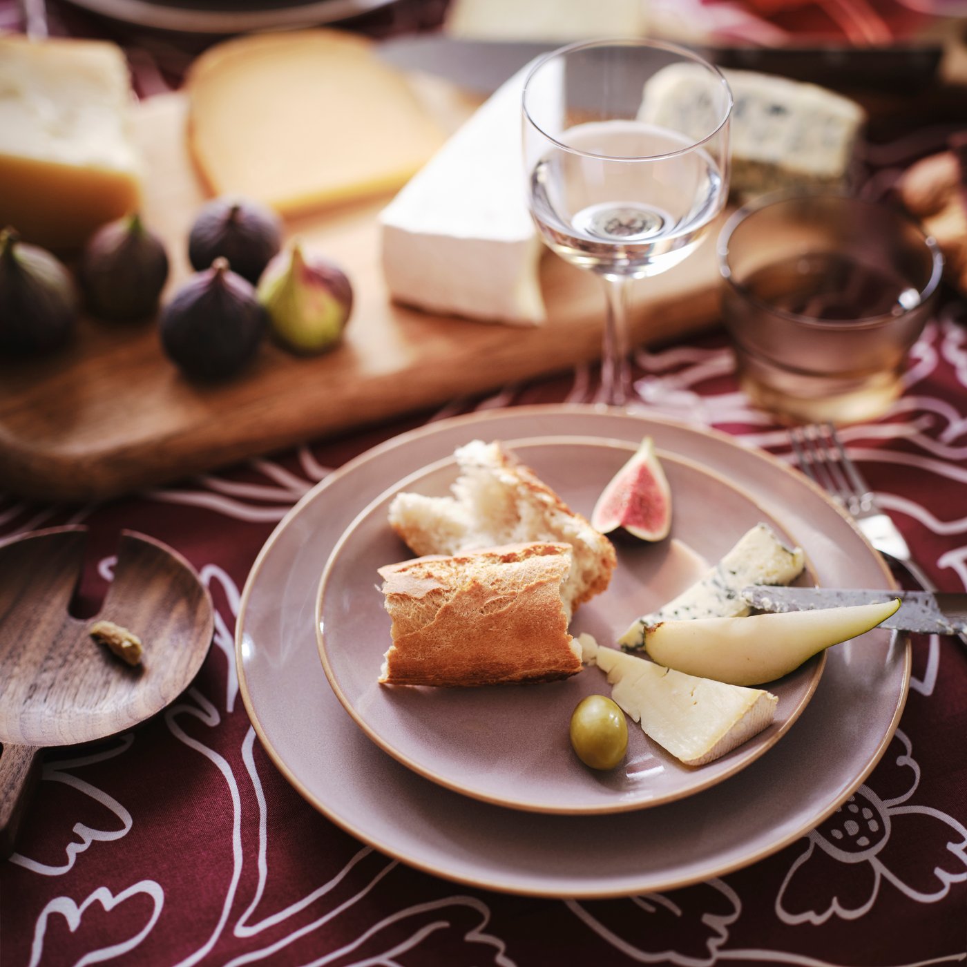 A table set with cheese and wine on a patterned tablecloth with mouth-blown STOCKHOLM wine glasses and FÄRGKLAR plates.