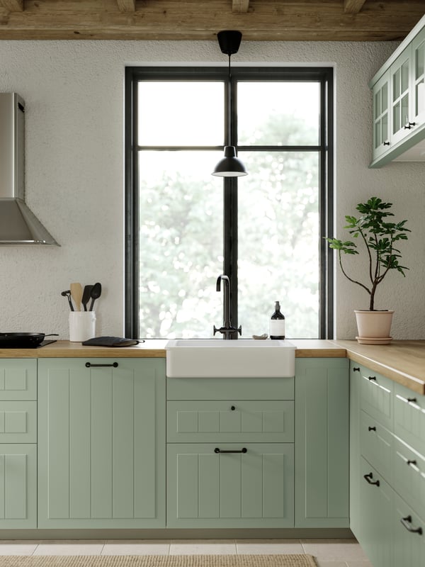 A kitchen with STENSUND kitchen fronts in light green with a window, kitchen appliances and a SKURUP pendant lamp in black.