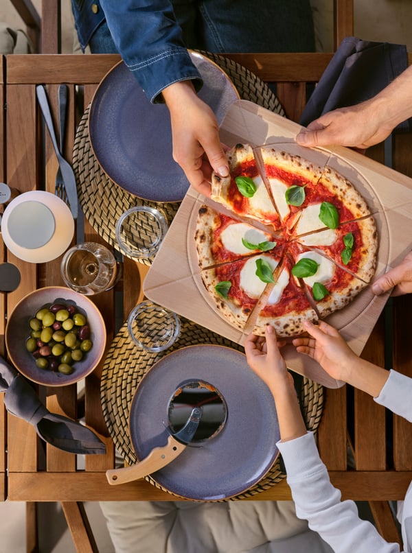 The hands of three people sharing a pizza on a SÅPÖRTMAL chopping board at an outdoor table made with FÄRGKLAR dinnerware.