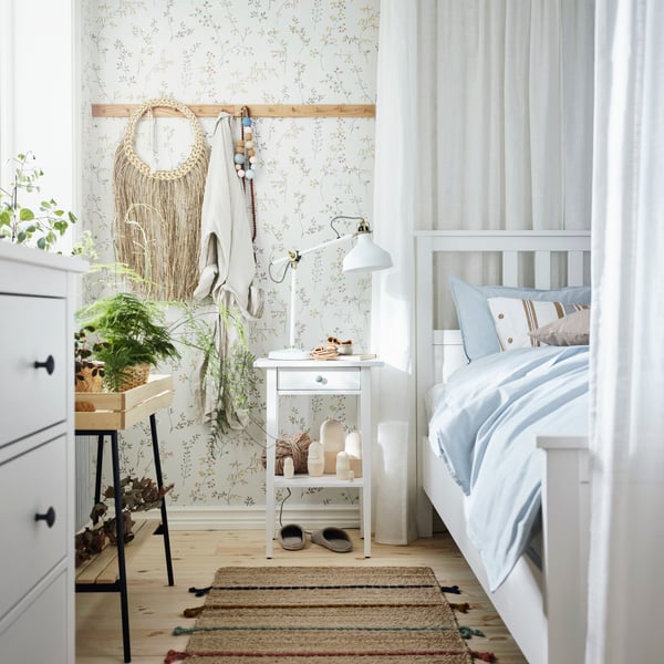 Ljust sovrum med vit säng, blommig tapet och en smal handvävd matta i naturtoner placerad bredvid sängen.