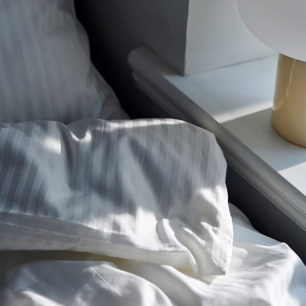The corner of a folded-over duvet covered in NATTJASMIN bed linen in white in a sunny space by a shelf.