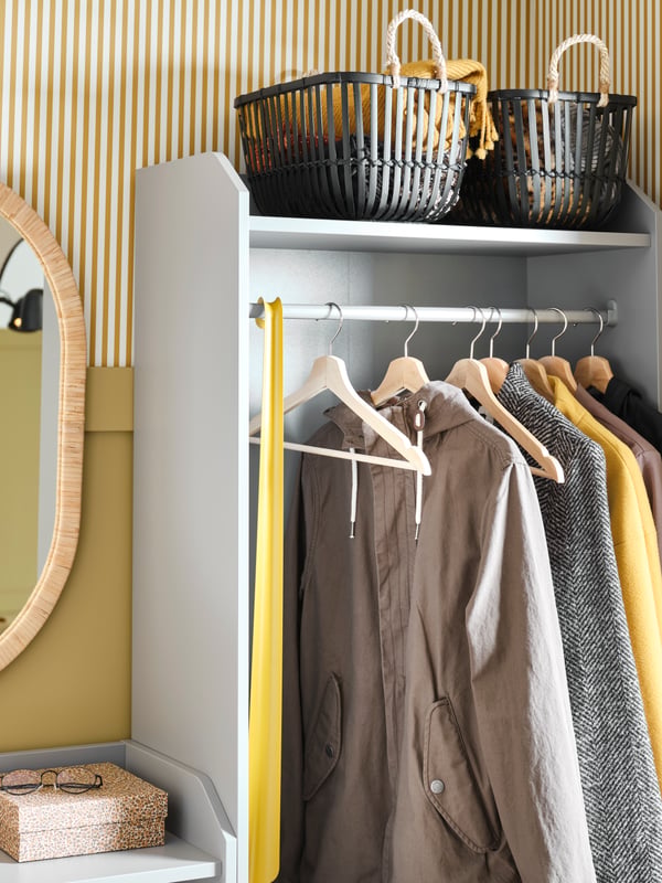 The top part of a HAUGA open wardrobe with jackets hanging on wooden BUMERANG hangers and LUSTIGKURRE baskets on top.