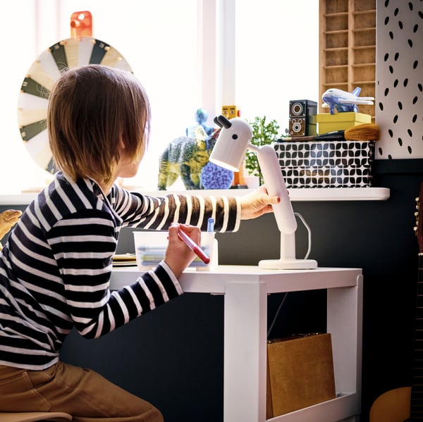Un jeune garçon assis, stylo à la main, à un bureau PÅHL blanc, ajustant une lampe de travail KRUX.
