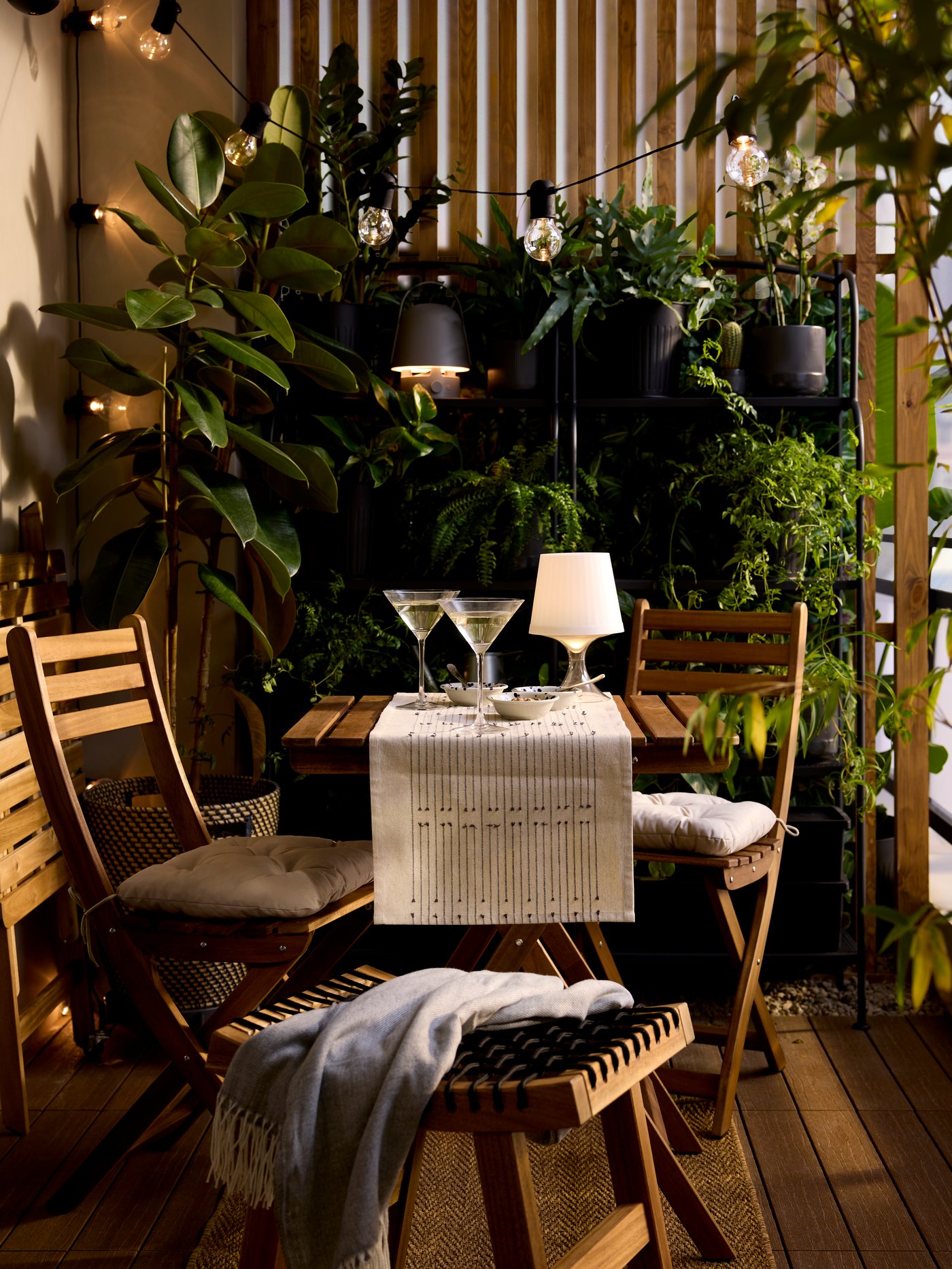 A plastic LAMPAN LED portable lamp with a white shade and clear base is lit, on the table of a bistro set in a lush balcony.