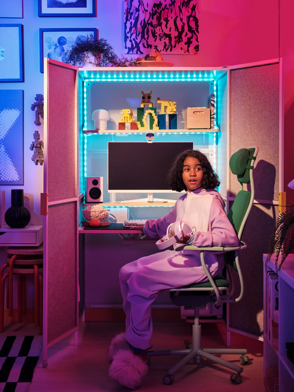 A young person sits at a BRÄNNBOLL gaming station that’s open and glows blue from multicolour VATTENSTEN LED lighting strip.