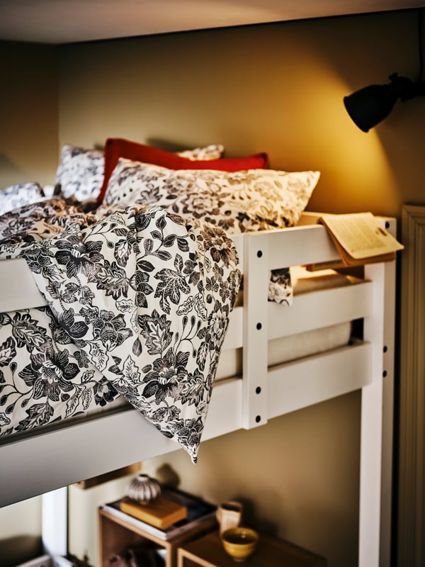 Ein STORÅ Hochbett mit der SKUGGNÄVA Bettwäsche. Eine schwarze Wandleuchte spendet dem Bereich Licht.