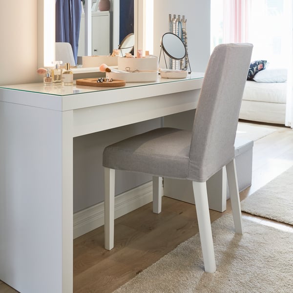 An IKEA BERGMUND upholstered dining chair with white legs and a light grey cover, in front of a white dressing table and lit mirror.