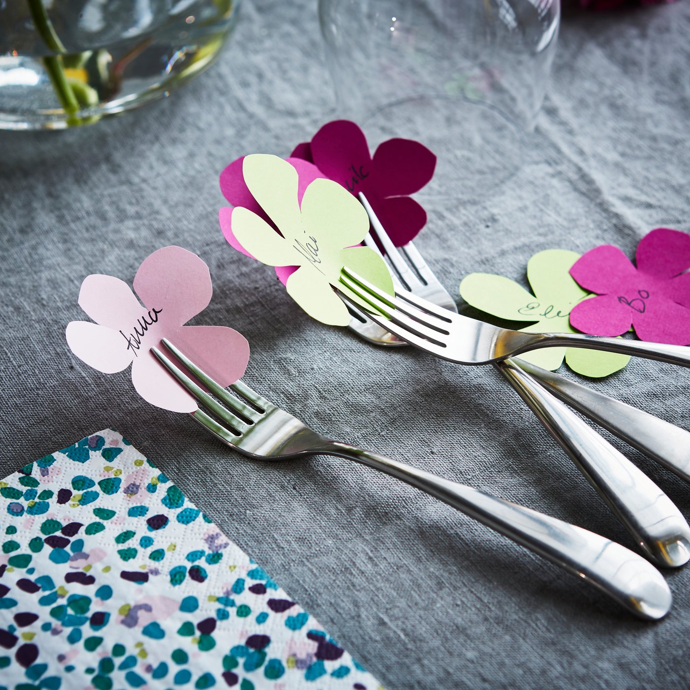 På et bord med en grå dug ligger gaffler med små blomsterformede kartonudklip, hvor der står et navn på.