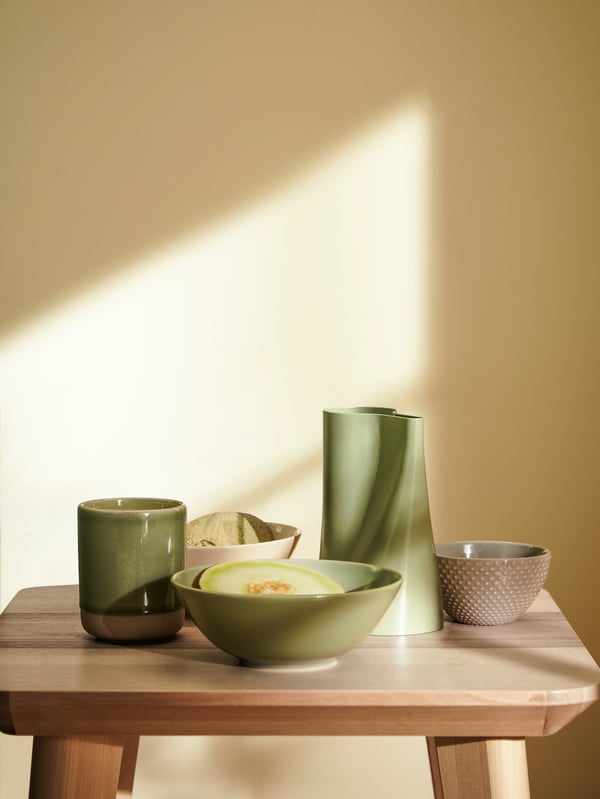 A wooden side table holding green and beige decorative objects including a vase, scented candle and bowls in ceramics.