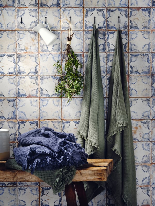 HÖGAFFEL wall hooks holding VALLASÅN bath towels in light green and a VALLASÅN bath towel in dark blue on a wooden bench.