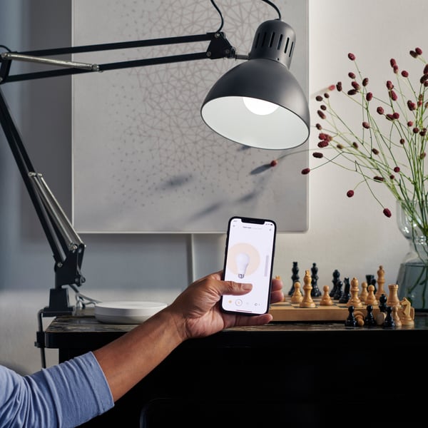 A person controls a lamp via the IKEA Home Smart app on a smart phone. A DIRIGERA hub is displayed on a table behind.
