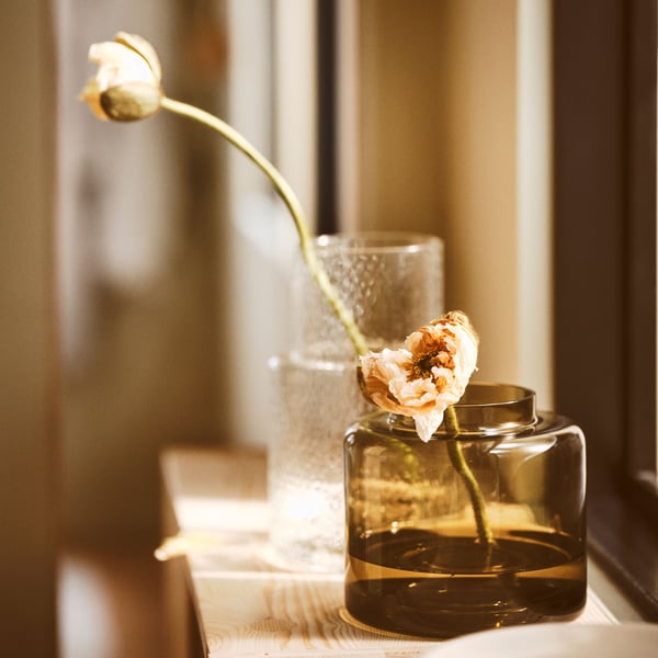 In a light-filled window are a green-brown KONSTFULL glass vase with a pink flower and a larger vase in clear glass.