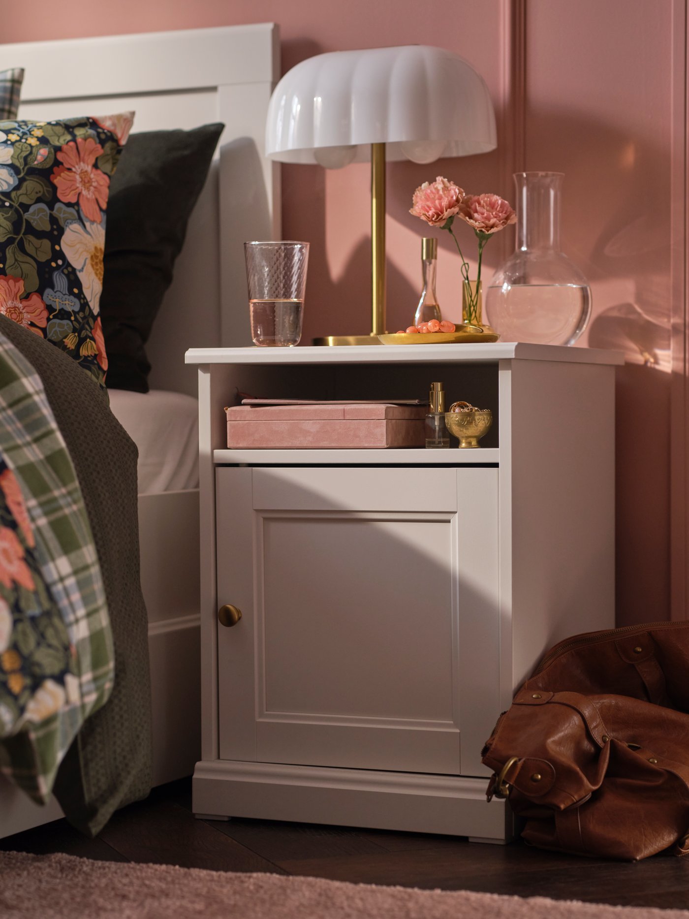 A FÅGELFJÄLLET side table in off-white with a white table lamp stands in a pink bedroom next to a bed with floral bedding.