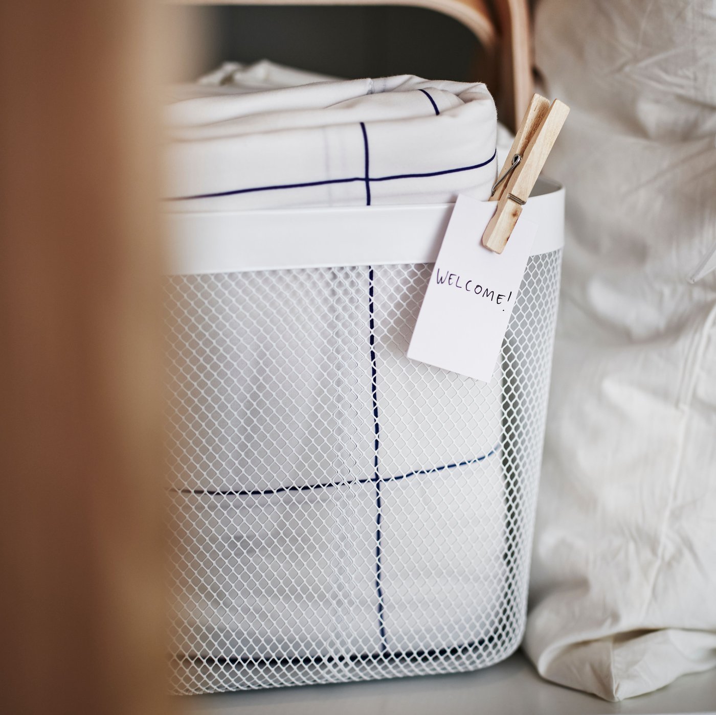 A white RISATORP basket with a label on the front, on a shelf holding folded white and blue check BREDVECKLARE bed linen.