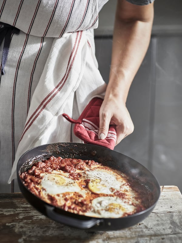 A person in a striped HILDEGUN apron is lifting a frying pan filled with eggs and red beans using a red HILDEGUN pot holder.
