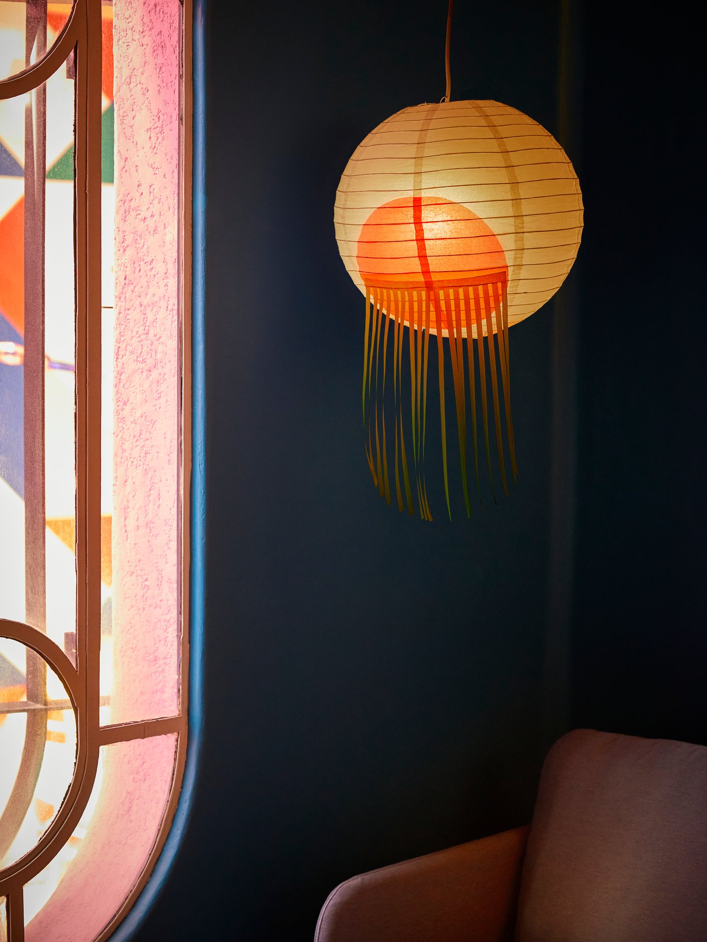 A white/pink, round ÖMSESIDIG pendant lamp shade in paper and powder coated steel hanging over an armchair next to a window.