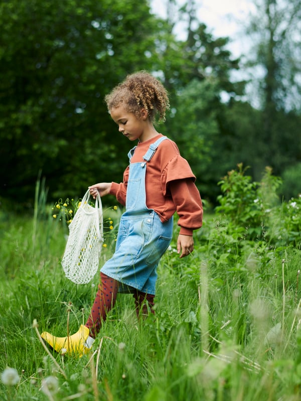 Fillette chaussée de bottes de pluie jaunes tenant un sac filet KUNGSFORS et traversant une prairie verte et arborée.