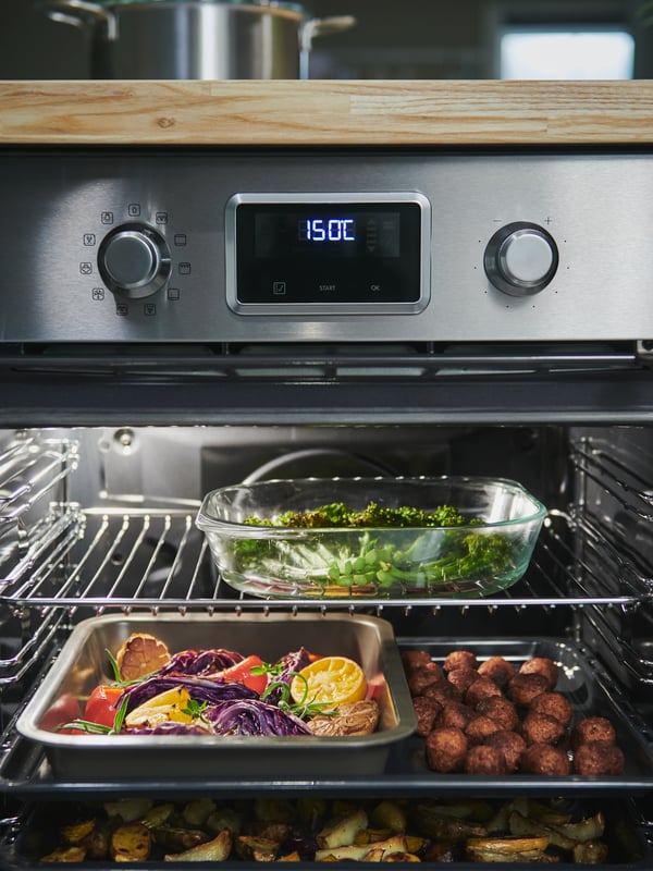 Un forno SMAKSAK in acciaio inox aperto con una teglia da forno FÖLJSAM in vetro trasparente, una teglia HEMMABAK e altri vassoi pieni di cibo - IKEA