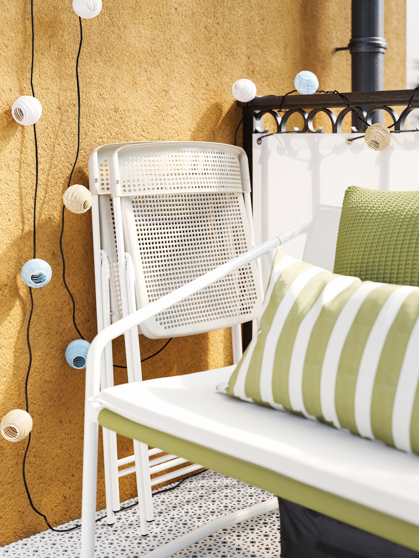 Deux chaises pliantes blanches/beiges contre un mur jaune, à côté d’une petite causeuse d’extérieur blanche décorée de deux coussins verts.