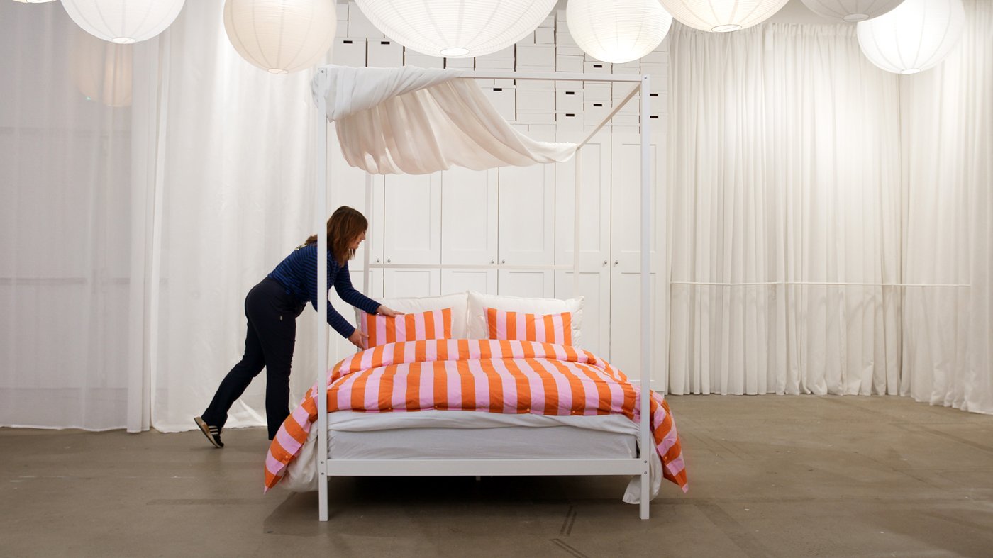 En IKEA inredare bäddar en säng med rosa- och orangerandiga sängkläder i ett ljust utrymme.